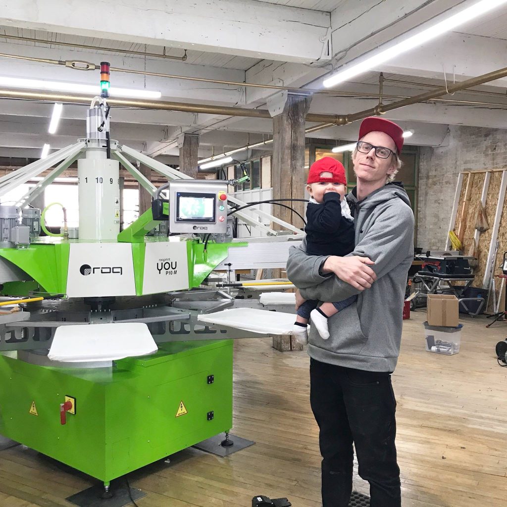 Someone holding a baby beside a green ROQ You automatic screen printing press on the shop floor