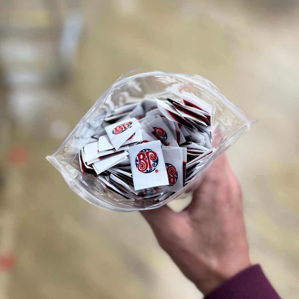 A hand holding a clear bag of white woven labels printed with a red and blue BP monogram