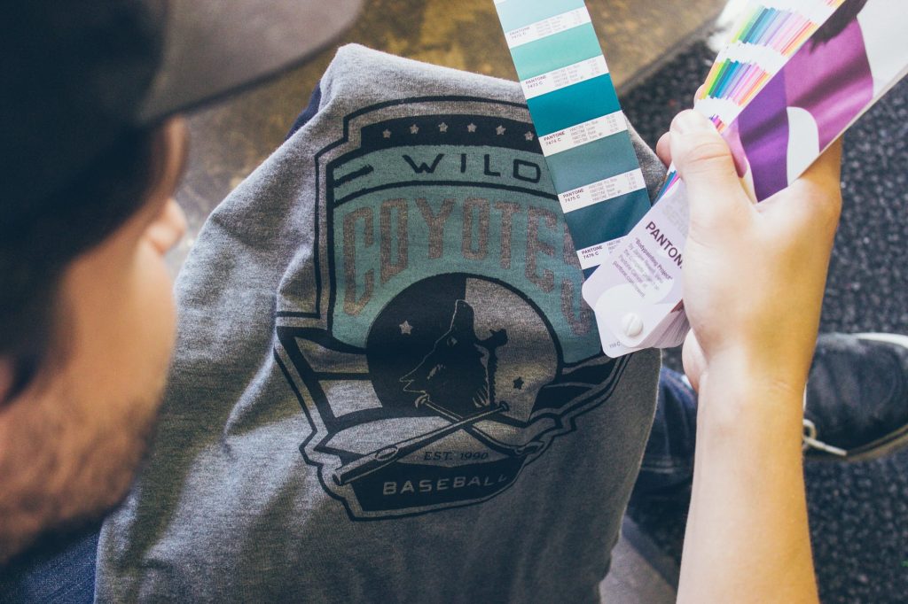 Holding Pantone colour fan decks over a grey tee printed with a Wild Coyotes Baseball crest, matching ink colours