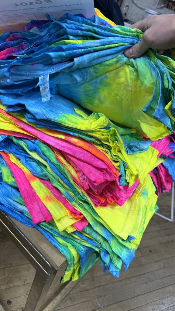 Stack of neon tie dye tees in blue, yellow and pink on a table, with a hand lifting the top one