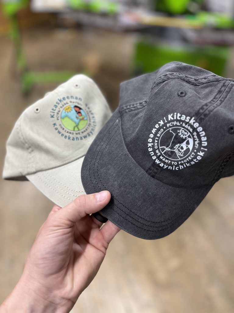 Two caps embroidered with the Kitaskeenan Kaweekanawaynichikatek logo, one stone and one charcoal
