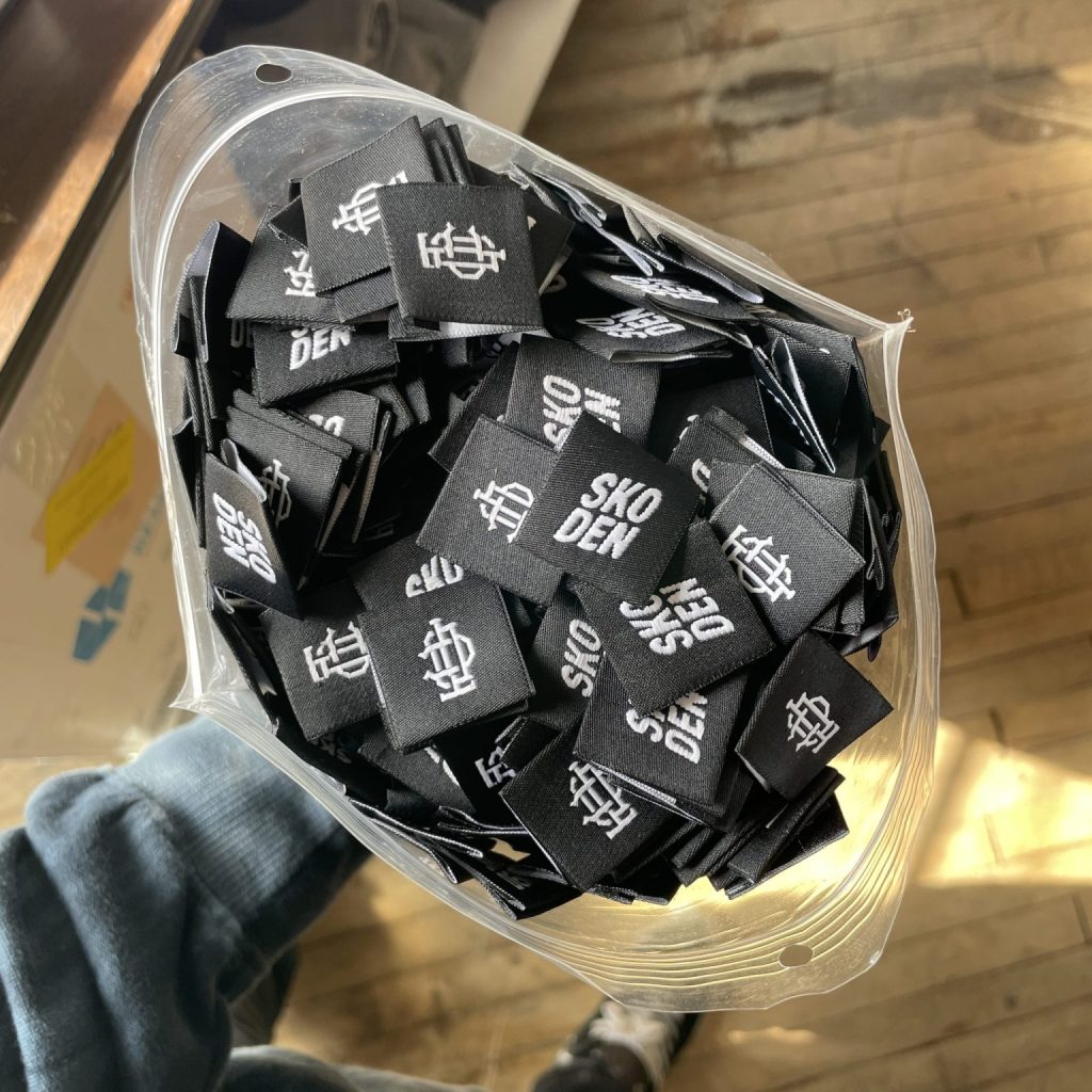 A clear bag of black woven labels stitched in white with Sko Den and a monogram, held over a wood floor