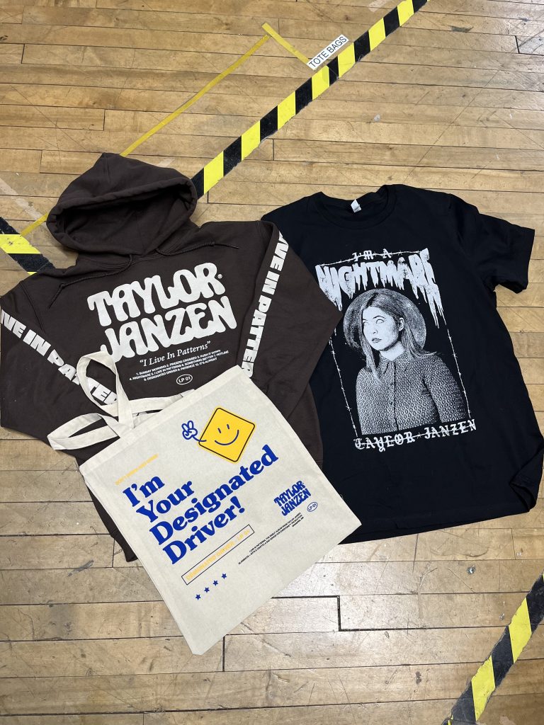 Taylor Janzen merch set, a brown hoodie, black tee and natural tote, laid out on the shop floor