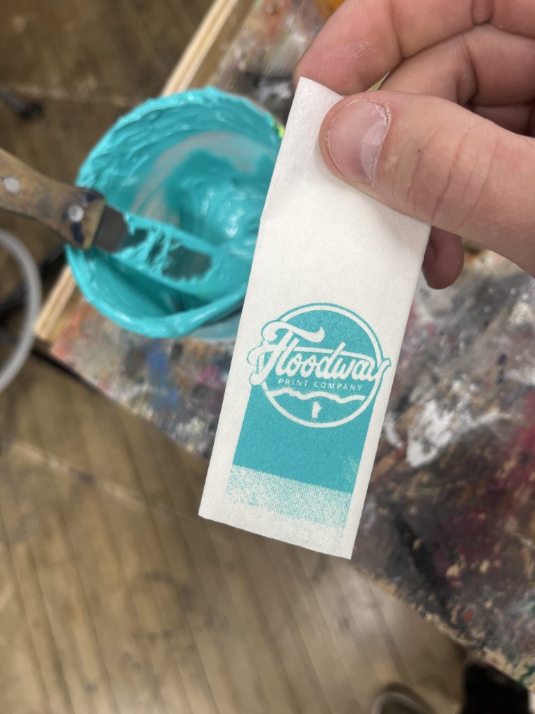 A hand holding a test print of the Floodway Print Company logo in teal ink, beside the ink tub
