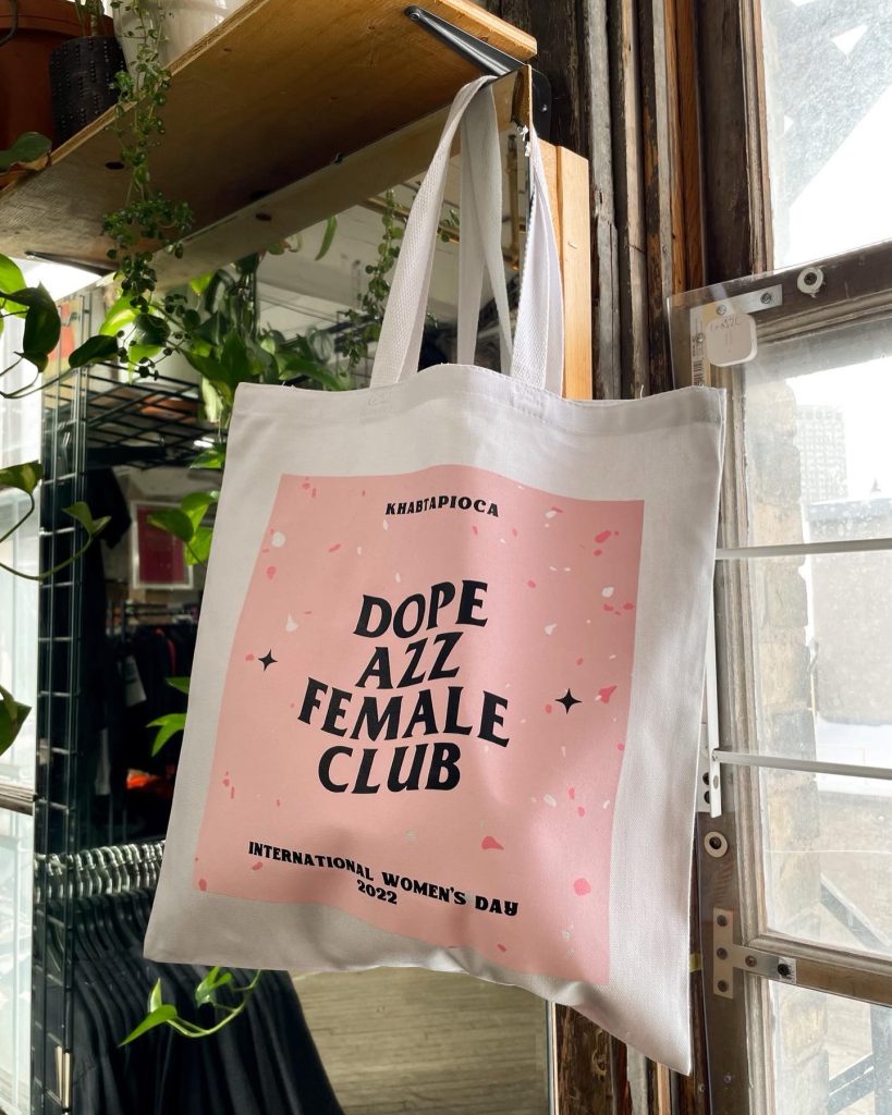 Canvas tote hanging in a window, printed with a pink panel reading Dope Azz Female Club, International Women's Day 2022
