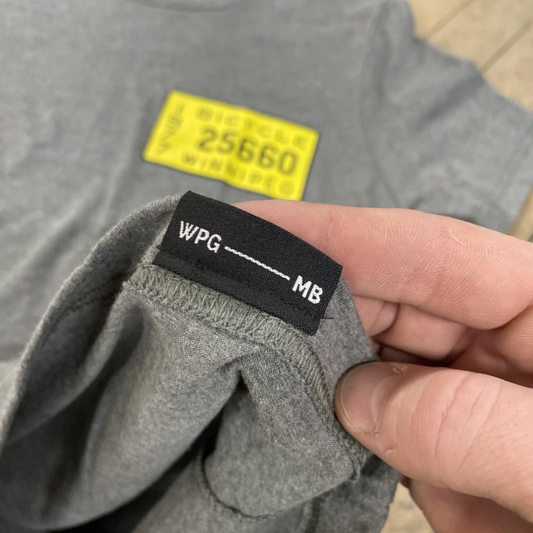 Grey tee with a custom WPG to MB woven neck tag and a yellow Winnipeg bicycle licence print