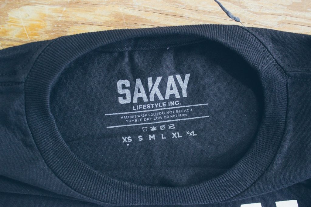 Inside back neck of a black tee printed with a SAKAY Lifestyle Inc. tag, care instructions and size list