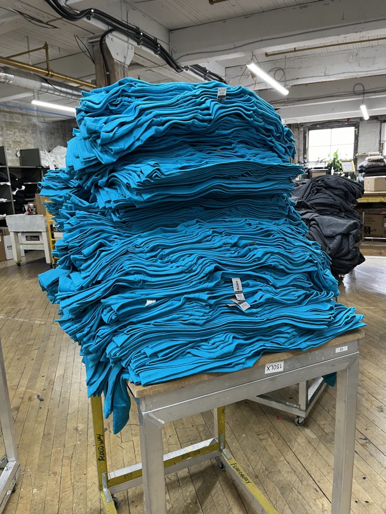 A tall stack of folded teal tees on a cart on the shop floor