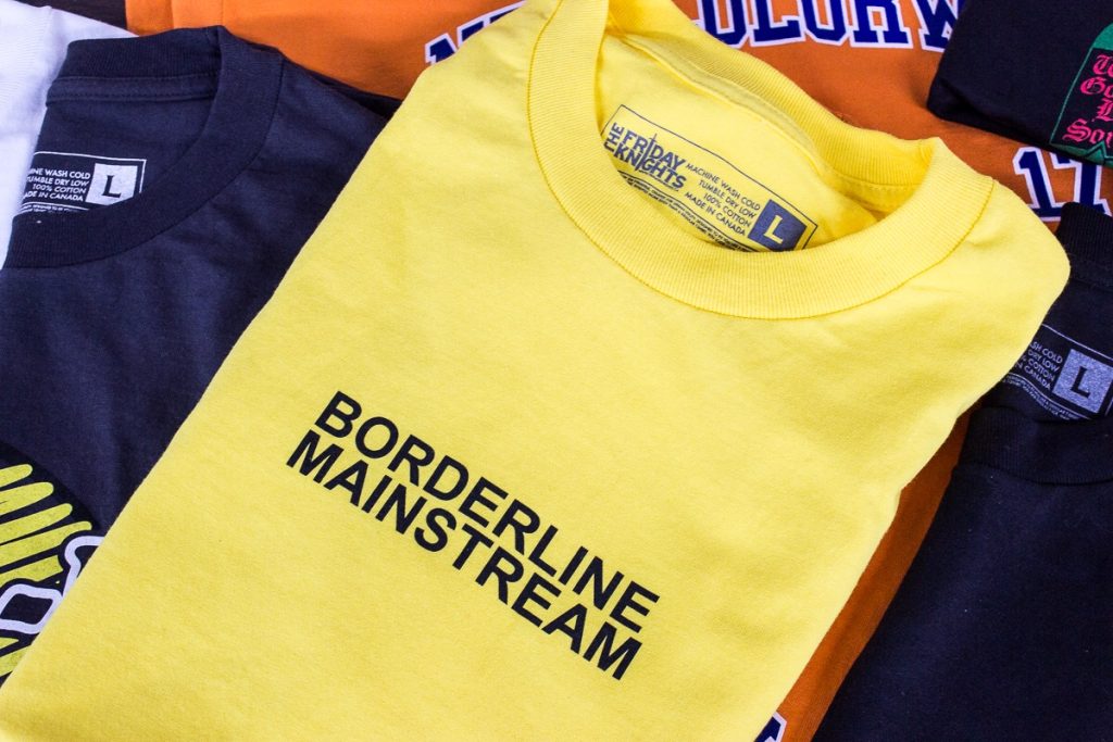Folded yellow tee printed Borderline Mainstream, stacked with navy and orange tees