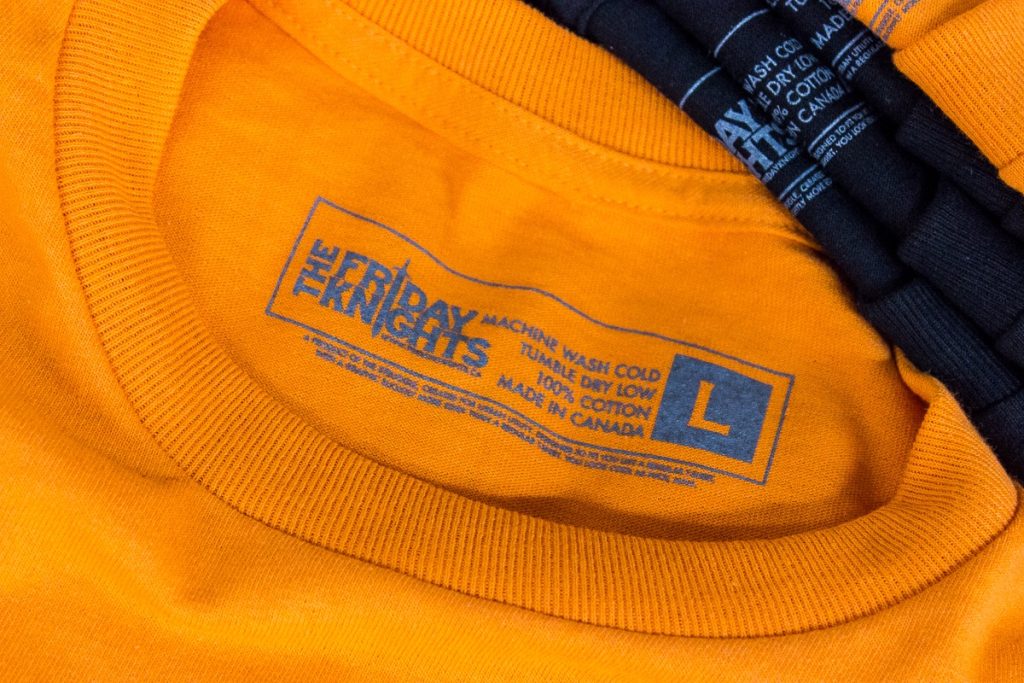 Close-up of a Friday Knights inside-neck tag print on an orange tee, with care details and size