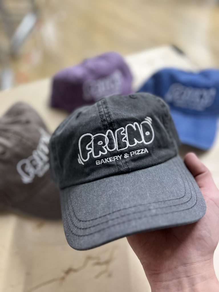 Two-tone grey cap embroidered Friend Bakery and Pizza, with purple, blue and brown caps behind