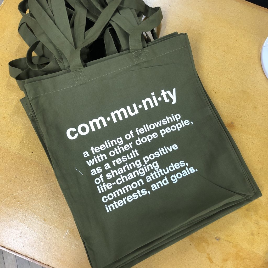 Stack of olive cotton tote bags printed with a white dictionary style definition of community