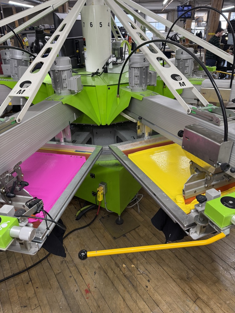 Automatic screen printing press mid-run, with pink and yellow ink flooded across the screens